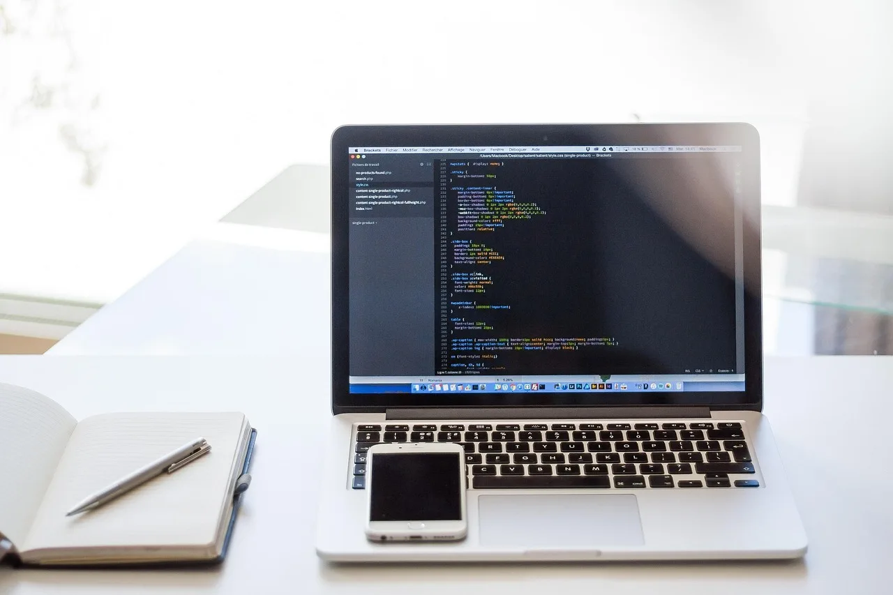 Laptop showing app code beside a smartphone and notebook, used for maintaining a mobile app after launch Laptop showing app code beside a smartphone and notebook, used for maintaining a mobile app after launch