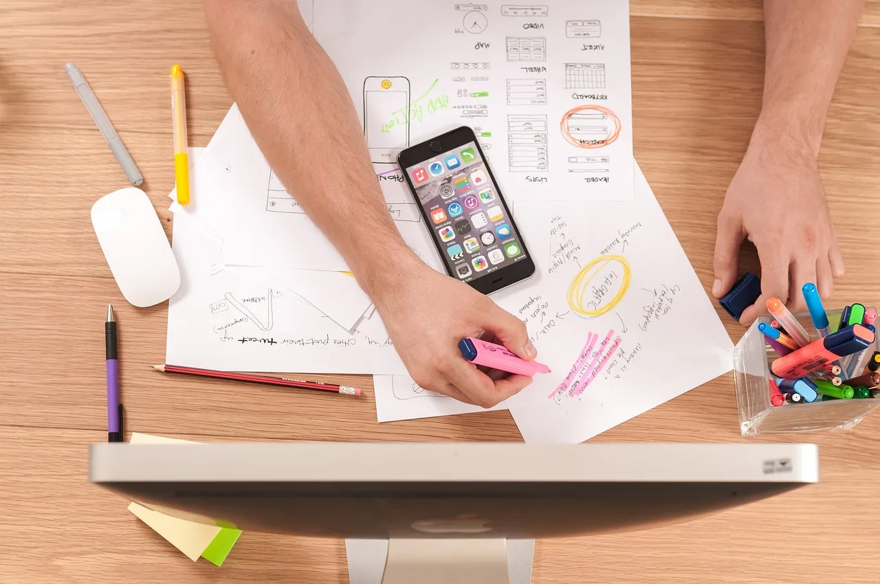 Designer sketching mobile app screens on paper next to a smartphone while planning an app build Designer sketching mobile app screens on paper next to a smartphone while planning an app build